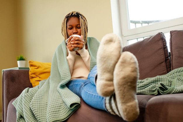 Afro woman wrapped on blue blanket and drinking hot tea in living room