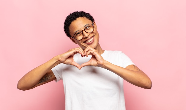 afro young black woman smiling and feeling happy, cute, romantic and in love, making heart shape with both hands