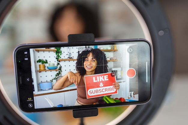 An AfroCaribbean woman showing a creative recipe with healthy fresh ingredients from her kitchen at home using her smart phone to film the cookery demonstration