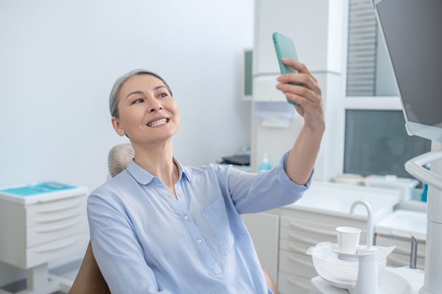 After teeth restoration. Smiling pretty mature woman with a smartphone at the dentists office