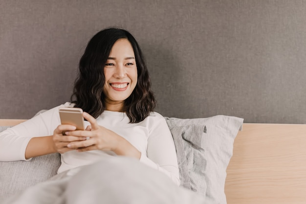 After wake up, Asian woman is smiling on the bed. She is looking in mobile phone and sending messages to her friends.