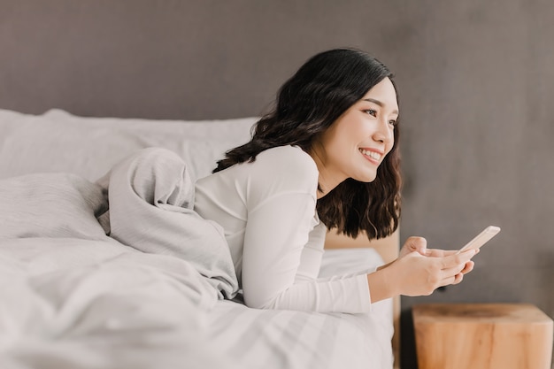 After wake up, Asian woman is smiling holding mobile phone on the bed. She is looking toward outside of her room.