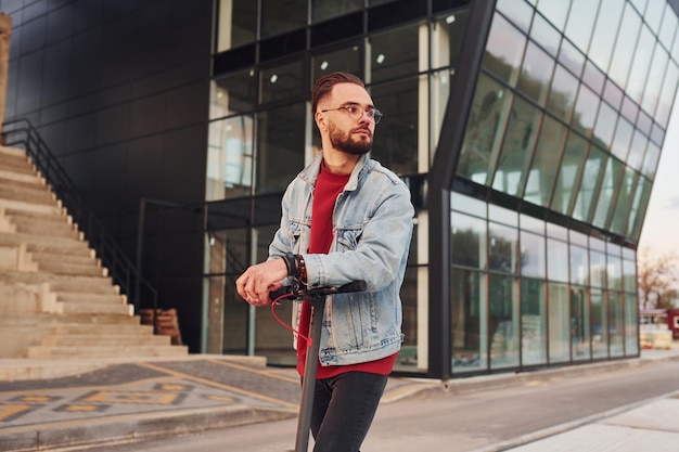 Against business building Handsome young guy in casual clothes standing with electric schooter outdoors at sunny daytime