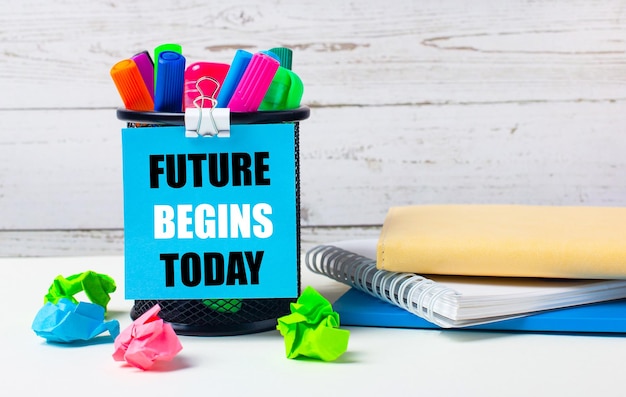 Against a light background of a wooden wall there is a glass with colored markers crumpled bright sheets of paper and a sheet of blue paper with the inscription FUTURE BEGINS TODAY