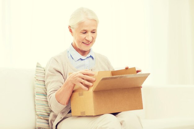 age, delivery, mail, shipping and people concept - happy smiling senior woman looking into open parcel box at home