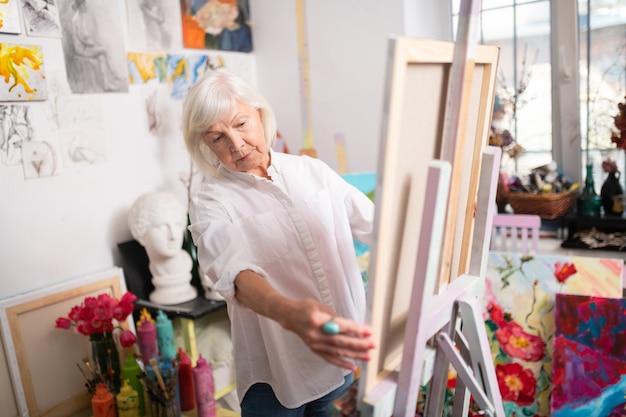 Aged artist. Aged artist with short blonde hair wearing white blouse standing near drawing easel