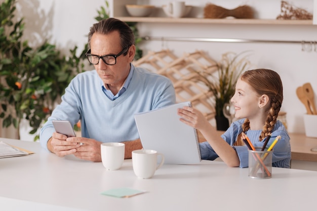 Aged busy concentrated grandfather using smartphone at home and ignoring granddaughter while working and texting sms