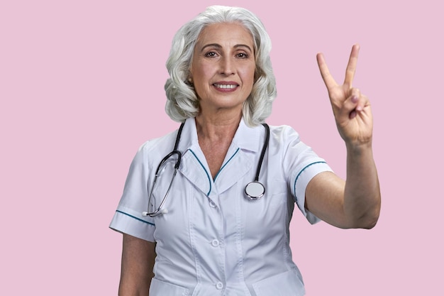 Aged caucasian female doctor show victory sign isolated on pink background