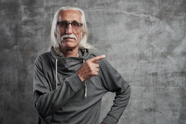 Aged gray haired Caucasian man points to the side with index finger, advertises something. Grandfather in glasses with mustache recommends buying your product