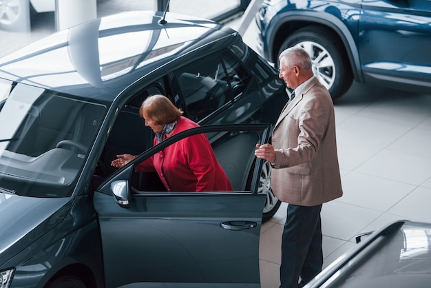 Aged man in formal wear supporting woman in choosing automobile.