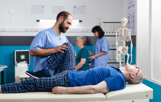 Aged man with leg muscle injury receiving chiropractic massage at kinetotherapy clinic. Assistant helping elder patient with knee pain, relieving pressure for physical recovery.