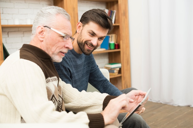 Aged man and young positive guy using tablet on settee