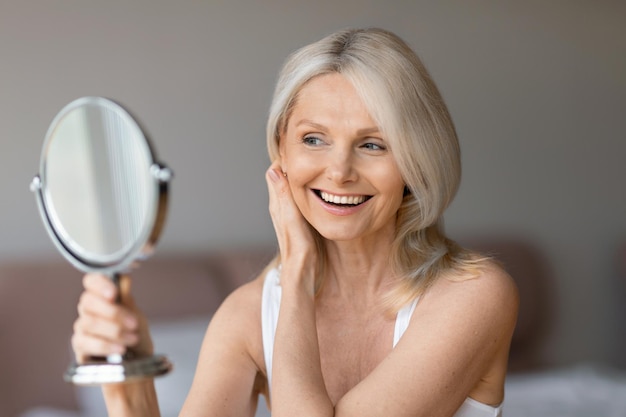 Aged natural beauty concept happy senior woman looking at mirror touching her face and smiling to