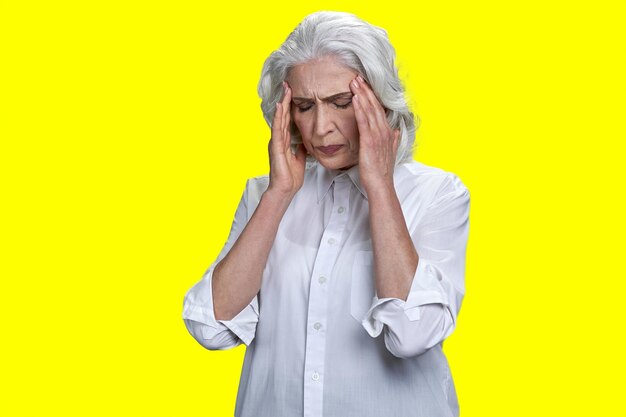 Aged woman holding head due to severe headache isolated on yellow backgrond