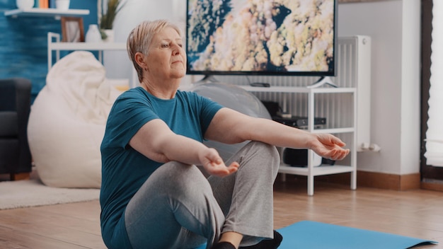 Aged woman preparing to meditate on yoga mat, with eyes closed and lotus position. Retired adult meditating at home after workout exercise, doing zen meditation for relaxation and calm.