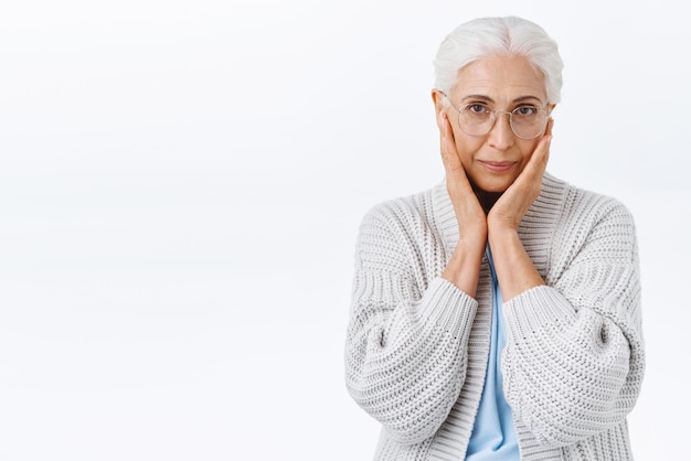 Aging holidays and people concept Charming senior lady with grey combed hair in glasses touch cheeks as blushing or dreaming smiling tenderly taking care facial wrinkles white background