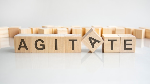 Agitate - word of wooden blocks with letters on a gray background. Reflection of the caption on the mirrored surface of the table. Selective focus.