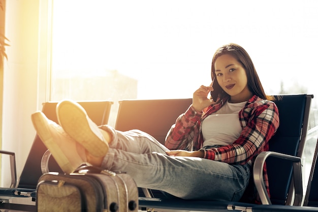 Air travel concept with young casual woman sitting with hand luggage suitcase. Airport woman talking on the phone at gate waiting in terminal.