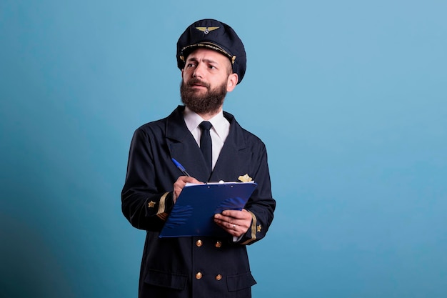 Airplane captain holding clipboard, filling documents, concentrated aviator with form. Aircraft pilot in aviation uniform writing in airport front view, studio medium shot on blue background