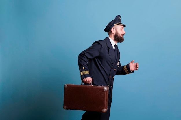 Airplane captain in professional airline uniform running late, carrying vintage suitcase. Pilot with luggage walking in airport, aviator holding baggage, side view medium shot