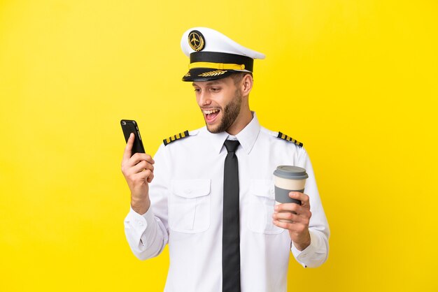 Airplane caucasian pilot isolated on yellow background holding coffee to take away and a mobile