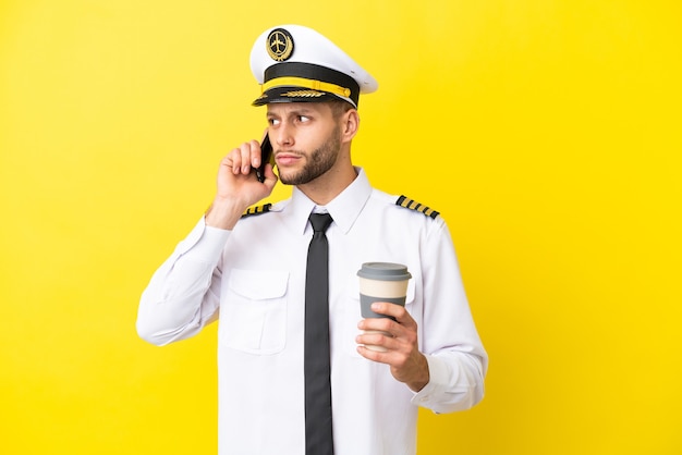 Airplane caucasian pilot isolated on yellow background holding coffee to take away and a mobile