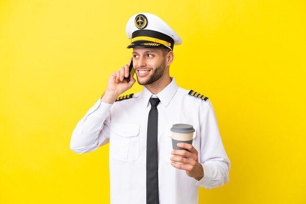 Airplane caucasian pilot isolated on yellow background holding coffee to take away and a mobile