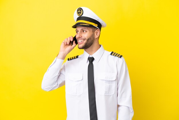 Airplane caucasian pilot isolated on yellow background keeping a conversation with the mobile phone