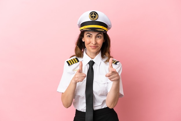 Airplane middle aged pilot woman isolated on pink background pointing to the front and smiling