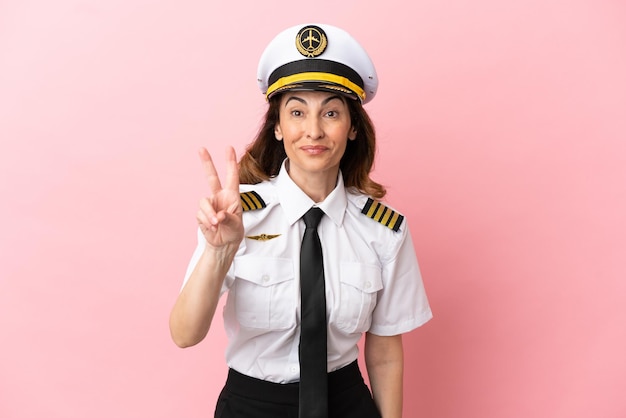 Airplane middle aged pilot woman isolated on pink background smiling and showing victory sign