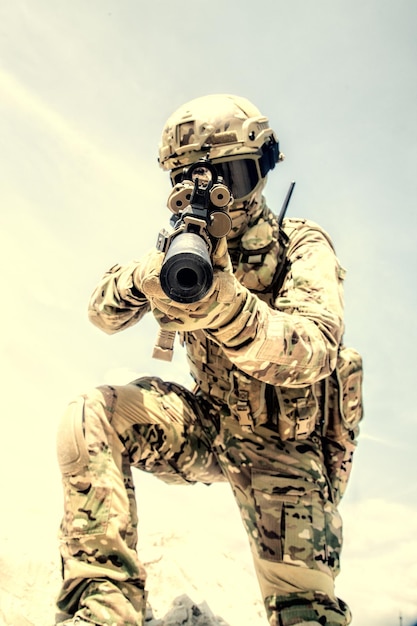 Airsoft war game player in army combat camouflage uniform, protected with helmet and tactical glasses, aiming in camera with optical sight on service assault rifle replica while climbing on sand dune