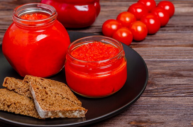 Ajvar or pindjur red vegetable spread pepper mousse from paprika and tomatoes in glass jars in a black plate on a rustic wooden table