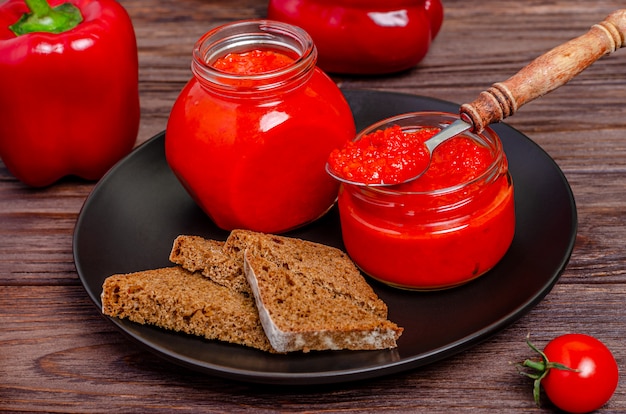 Ajvar or pindjur red vegetable spread or pepper paprika and tomato mousse in glass jars in a black plate on a rustic wooden table