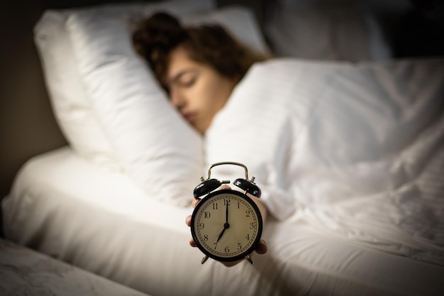 Alarm clock on the bed with sleeping woman behind Girl keeps alarm clock by her hand