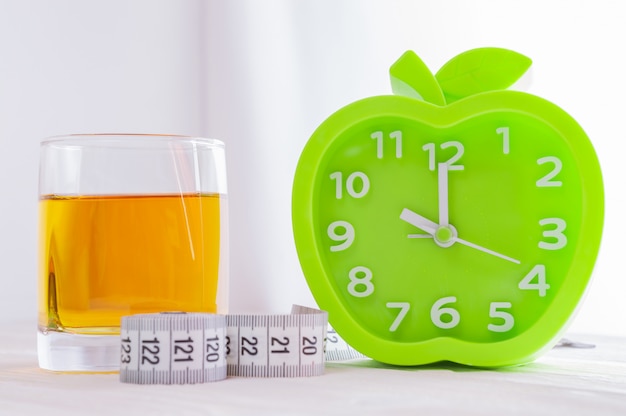 Alarm clock, a glass of juice and measuring tape on a white wooden table. 