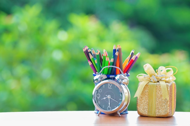 Alarm clock old retro style with color pencil and gift box on green burred background.