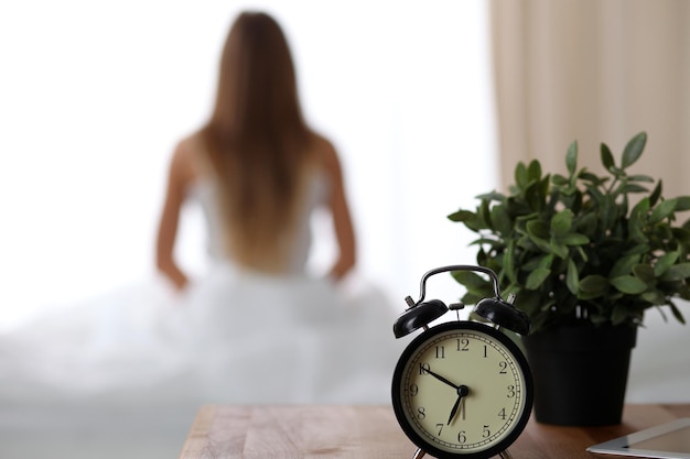 Alarm clock standing on bedside table has already rung early morning to wake up woman in bed sitting in background Early awakening not getting enough sleep oversleep time line concept