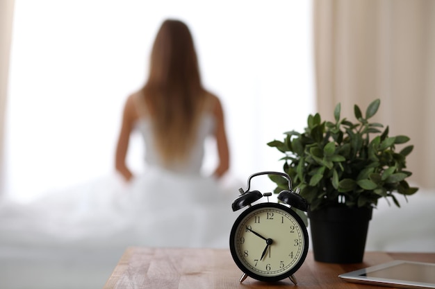 Alarm clock standing on bedside table has already rung early morning to wake up woman in bed sitting in background Early awakening not getting enough sleep oversleep time line concept