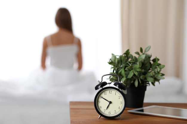 Alarm clock standing on bedside table has already rung early morning to wake up woman is stretching in bed in background Early awakening not getting enough sleep oversleep concept