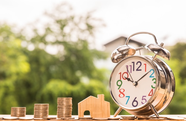 Alarm clock and wooden house model and step of coins stacks, nature background, money, saving and investment or family planning concept