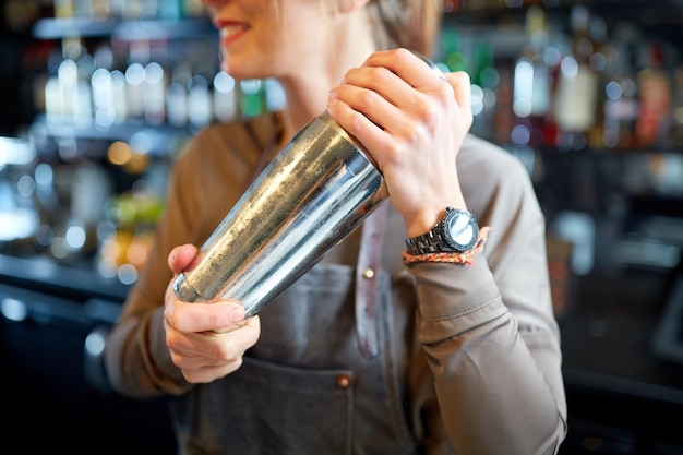 alcohol drinks, people and luxury concept - close up of smiling woman bartender with steel shaker preparing cocktail at bar