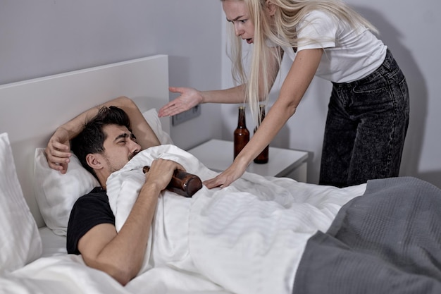 Alcoholic man lying on bed with alcohol while blonde wife is scolding him