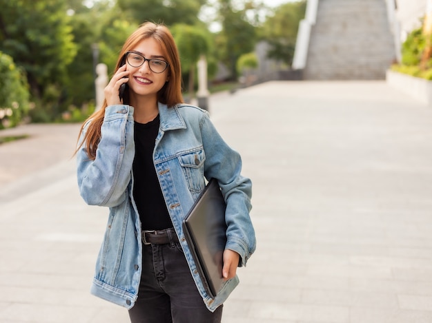 All in business. Young stylish woman in a denim jacket and glasses holding laptop and talking on phone while walking on the city.