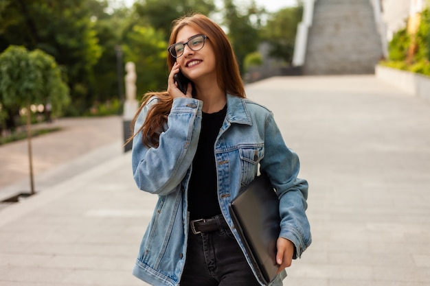 All in business. Young stylish woman in a denim jacket and glasses holding laptop and talking on phone while walking on the city.