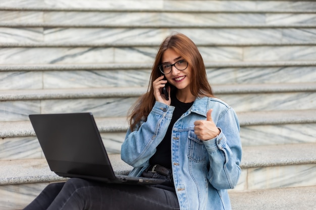 All in business. Young stylish woman in a denim jacket and glasses uses a laptop and talking on the phone while sitting on the stairs in the city. Remote work.