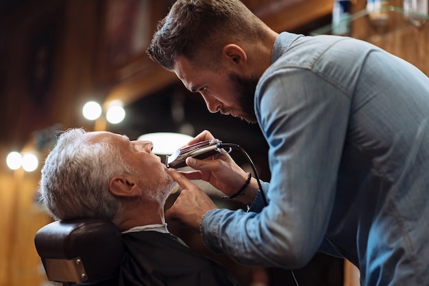 All details. Side view of young handsome bearded hairdresser trimming beard of senior barbershop client.