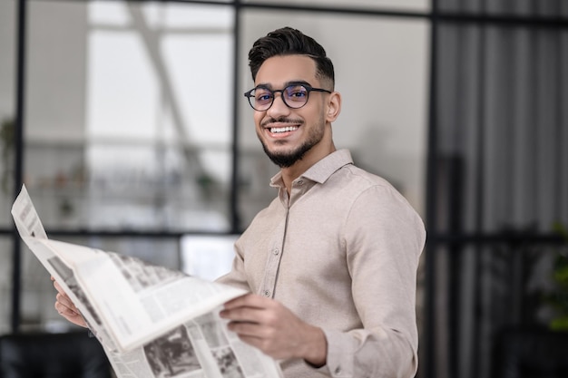 All excellent. Happy attractive young man in glasses with toothy smile holding newspaper looking at camera standing indoors