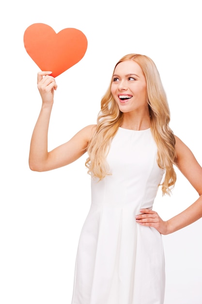 All thoughts about love. Beautiful young blond hair woman holding heart shaped valentine card and looking at it with smile while standing isolated on white