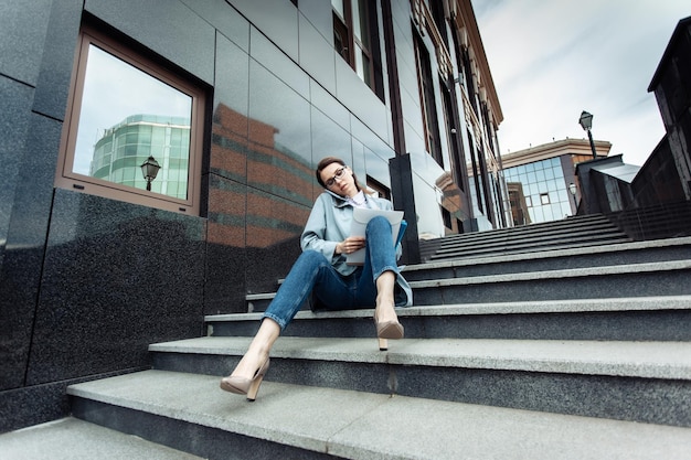 All at work modern business lady talking on the phone and writing important information on paper while sitting on the stairs