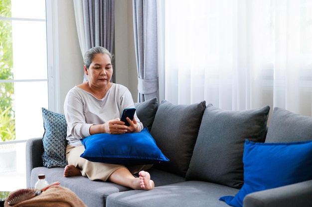 Alone retired asian older woman sitting on sofa and talk to her family by mobile phone is using internet at living room relax time at home